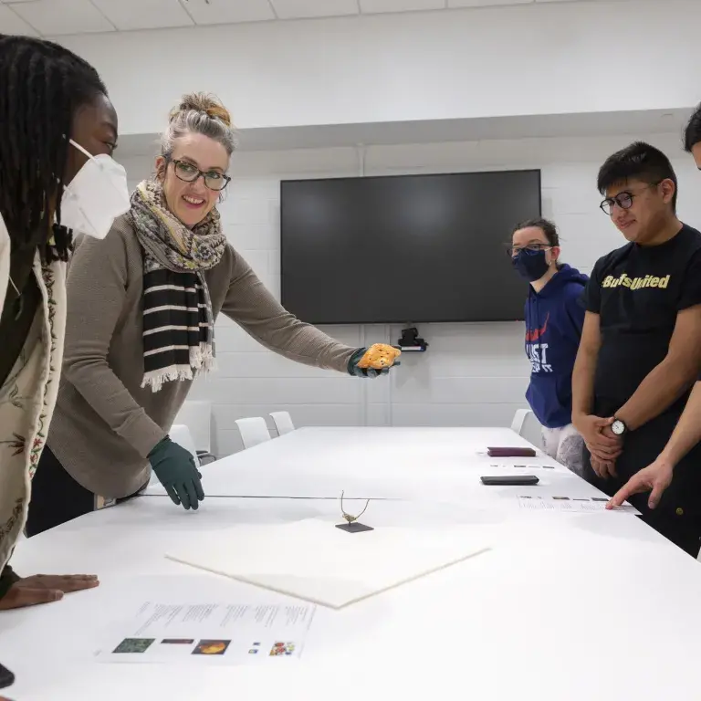 A woman shows a small artifact to a group of students around a table.